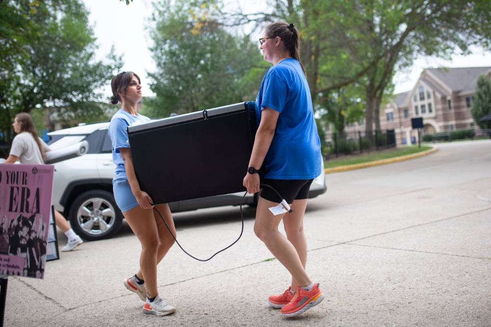 Two alums carrying a big box into a dorm.